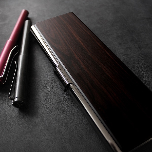 A wooden pen case (黒檀) with metal trim placed on a dark surface next to two pens.