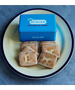 A blue ÉCHIRÉ patisserie tin positioned behind several iced square cookies on a plate.