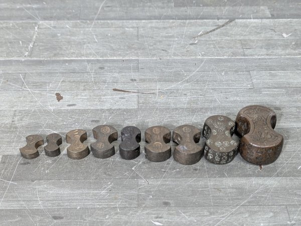 Nine iron 分銅 (traditional Japanese weights) of varying sizes arranged in a row on a wooden surface.