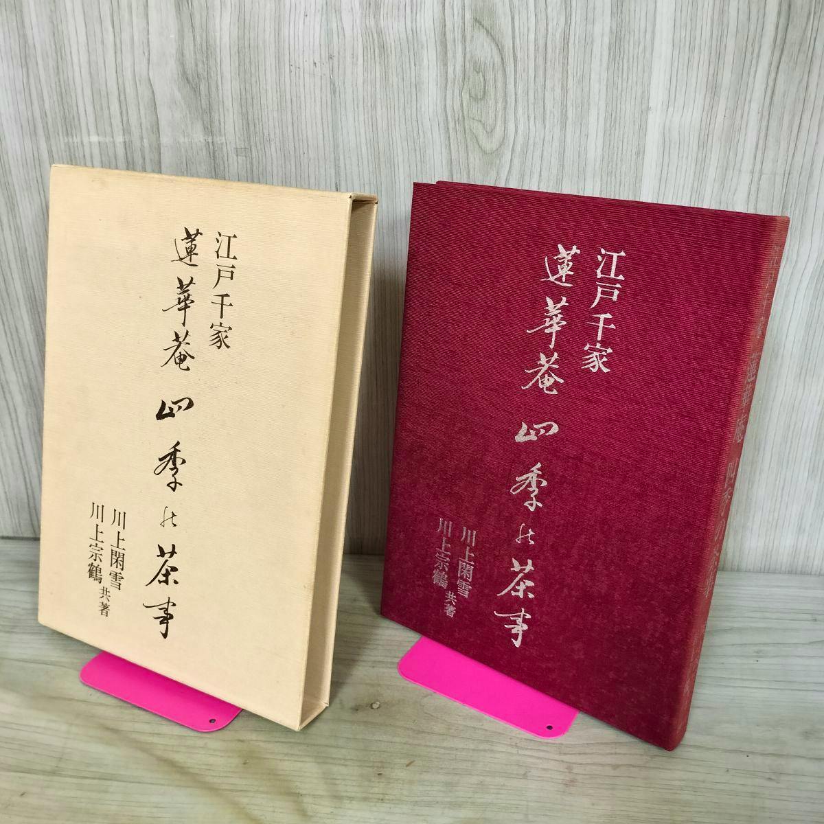 A red hardcover book and its beige slipcase standing side-by-side on pink supports against a wood background.