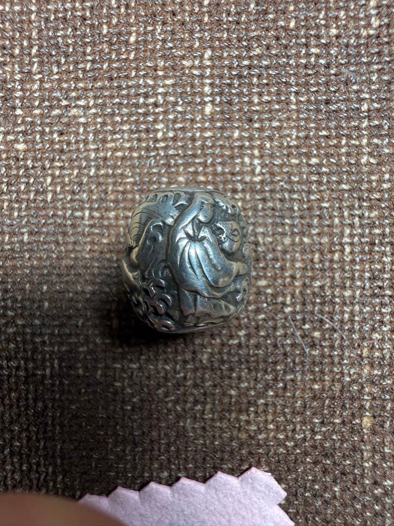 Silver personal ornament bead showing an engraved Kannon figure placed on brown woven fabric.