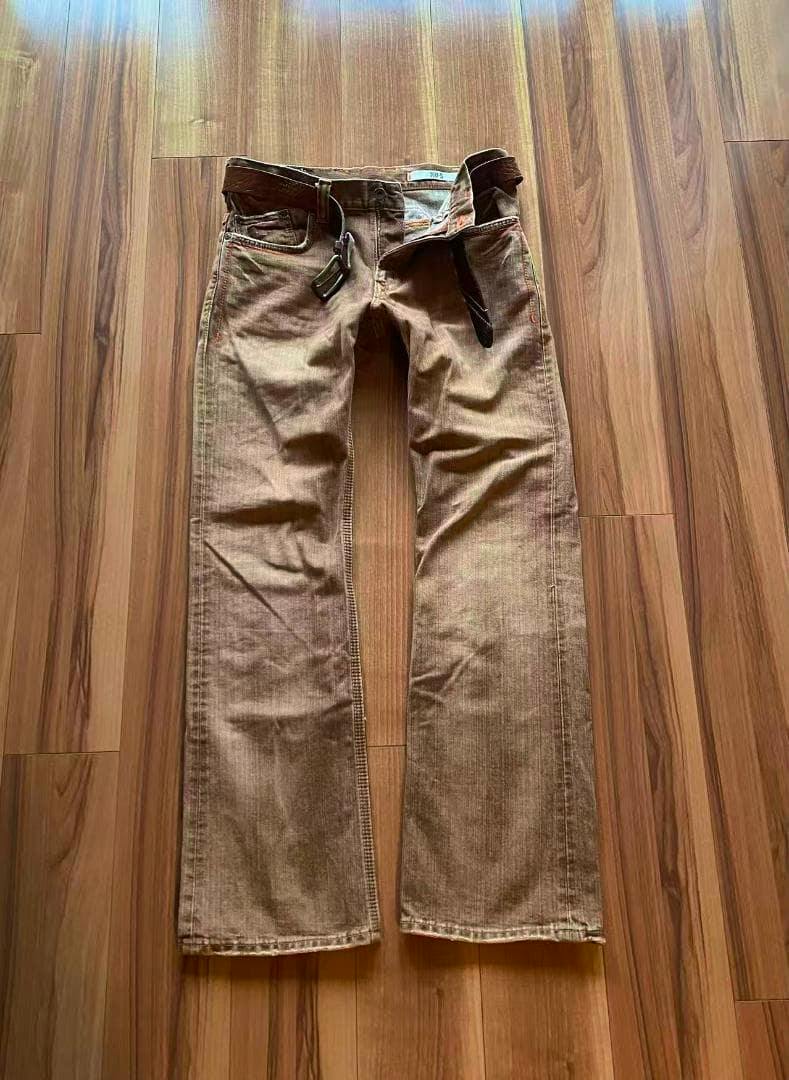 Brown bootcut denim pants laid flat on a wood floor with a belt threaded through the waistband.