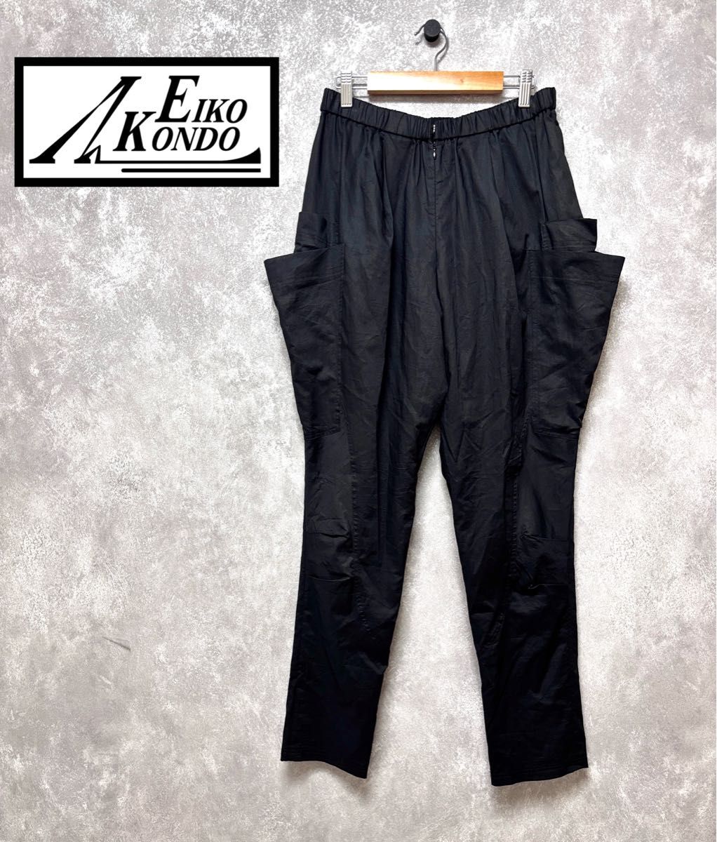 Black women's cotton pants with large side pockets hanging on a wooden clip hanger against a grey textured background with an Eiko Kondo logo in the corner.