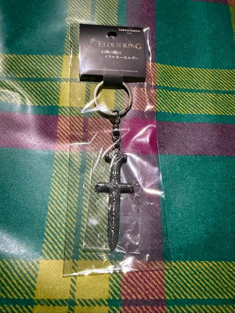 Packaged metal bag charm keychain in the shape of the stone sword from Elden Ring, shown on a plaid fabric background.