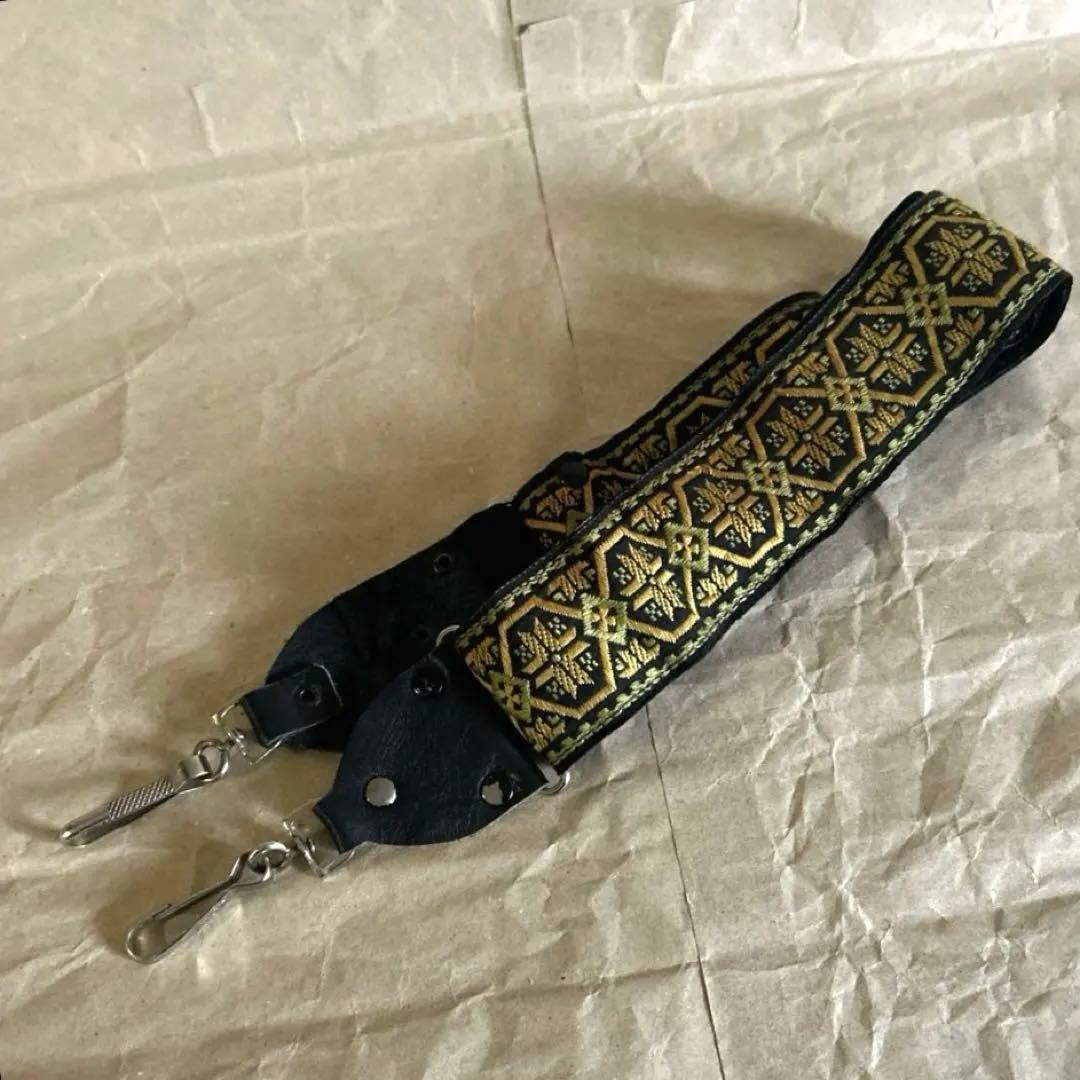 Embroidered vintage camera shoulder strap with patterned chirilian tape, leather ends, and metal clips lying on paper.