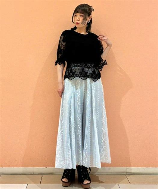 Model wearing light-colored embroidered flare pants and a black lace top, standing against a peach wall.
