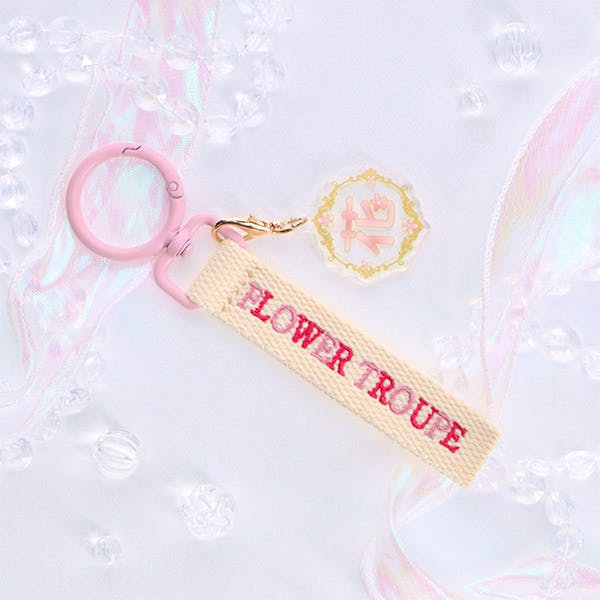 Beige embroidered fabric keychain tag reading 'FLOWER TROUPE' attached to a pink key ring and a small decorative charm.