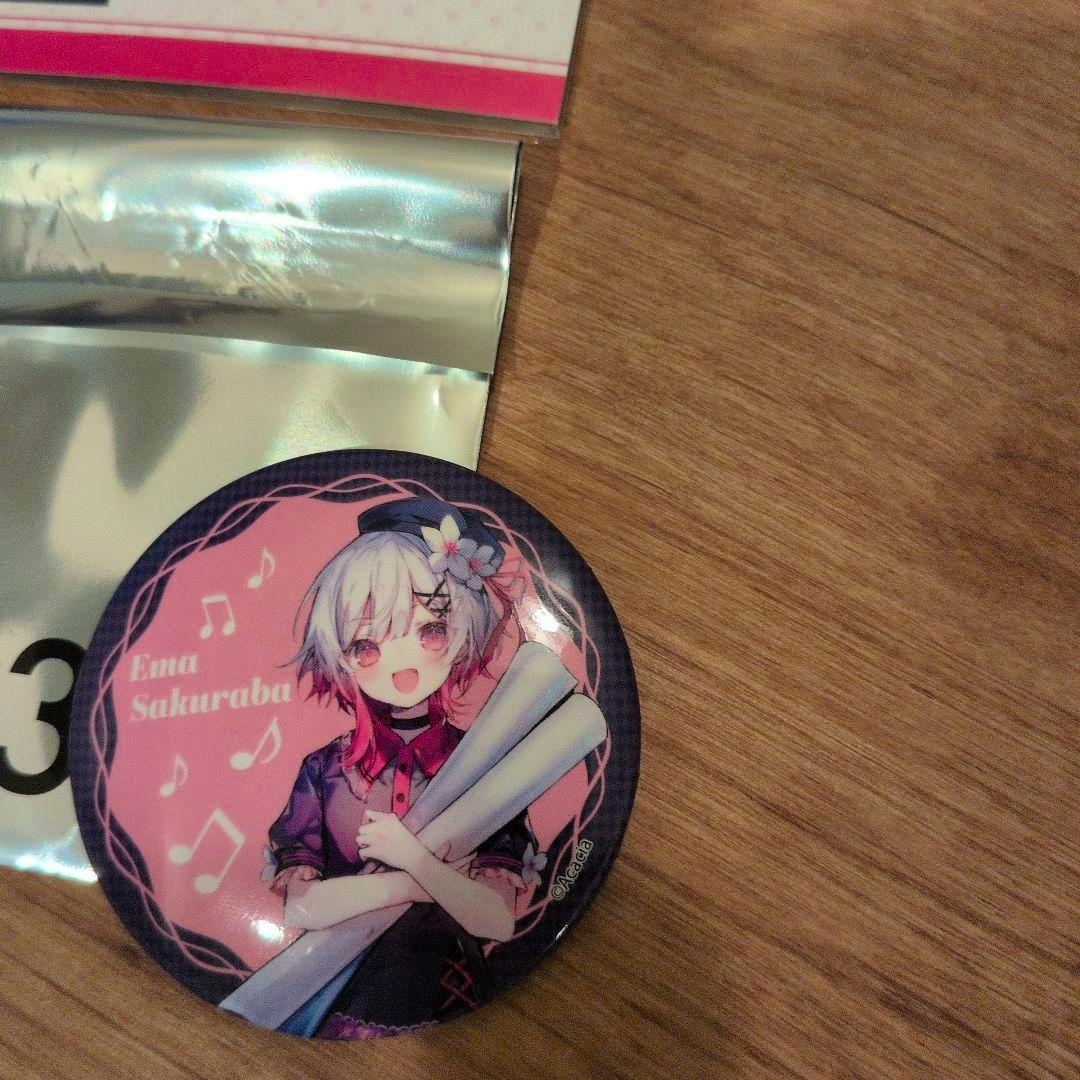 Close-up photo of a round pink can badge showing the character Ema Sakuraba with white hair, musical notes, and crossed arms holding rolled papers on a wooden surface.