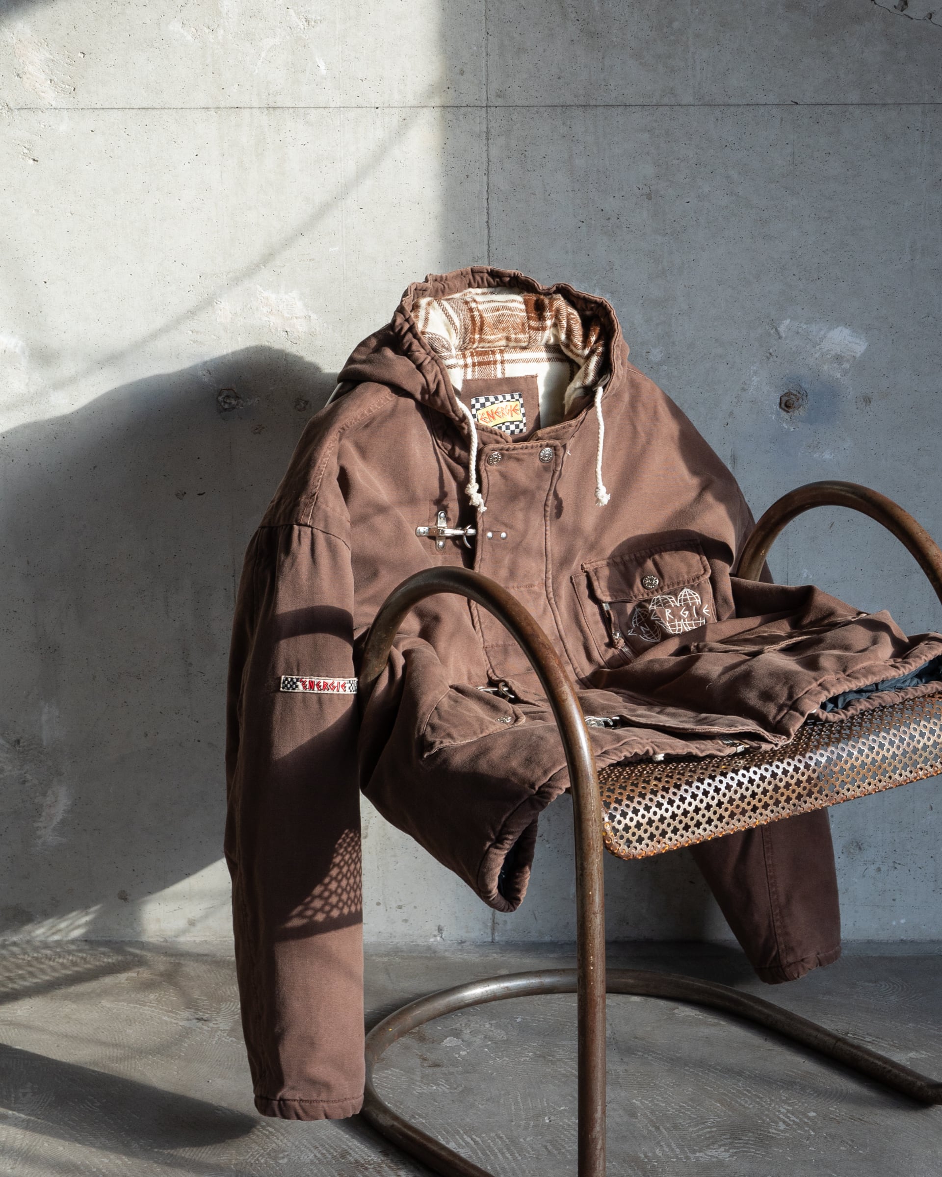 Brown ENERGIE duck fireman-hook jacket with flannel check lining and quilted body draped over a chair against a concrete wall.