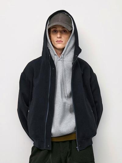 A person wearing a black melton parka with the hood up over a gray hoodie and a baseball cap.