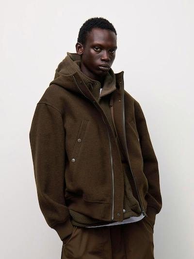 A model wearing the brown COZY MELTON hooded parka sweater with a front zipper and snap-pocket details.