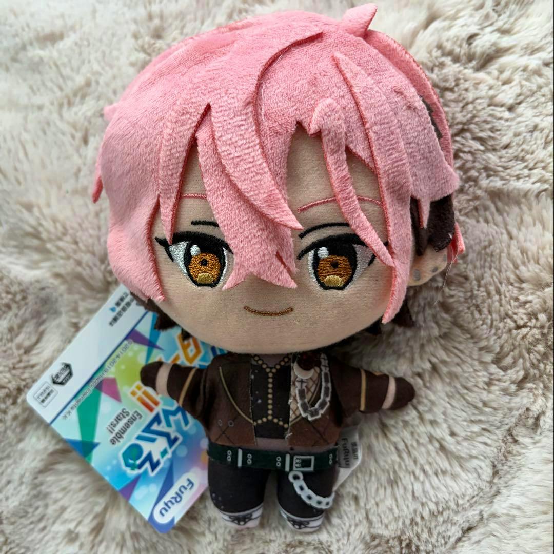Front view of the plush toy 久遠舞珠 (あんさんぶるスターズ) with pink hair, embroidered face and outfit, and an attached product tag on a beige fuzzy surface.
