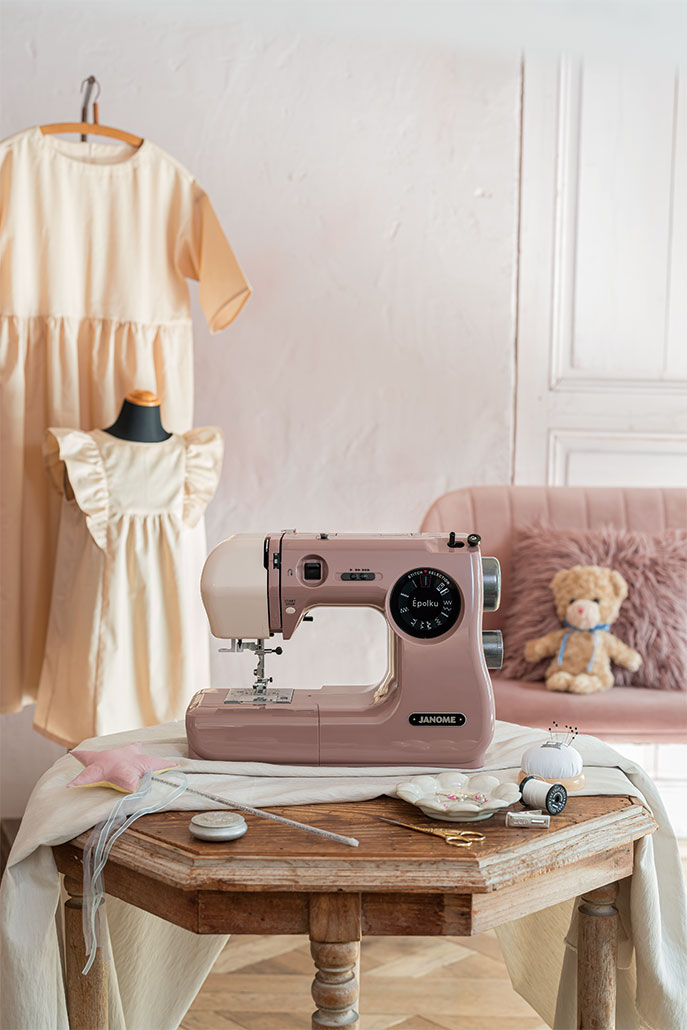 Pink handheld sewing machine on a wooden table with a dress on a mannequin and a teddy bear in the background.