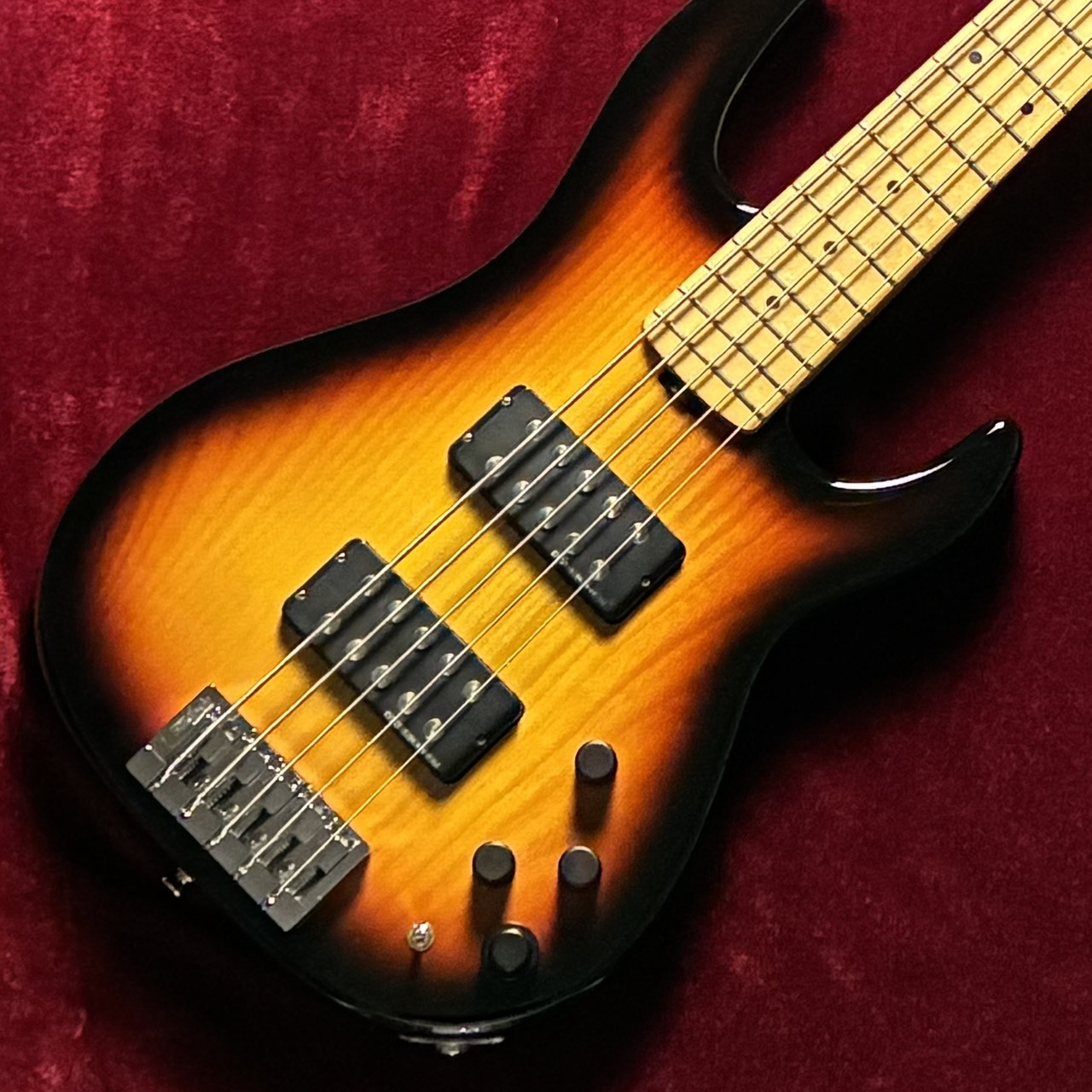 Close-up photo of a five-string bass guitar body on a red background showing a sunburst finish, neck, two pickups, bridge, and control knobs.