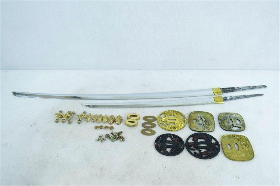 Two imitation sword blades and assorted metal fittings and tsuba pieces laid out on a light background.