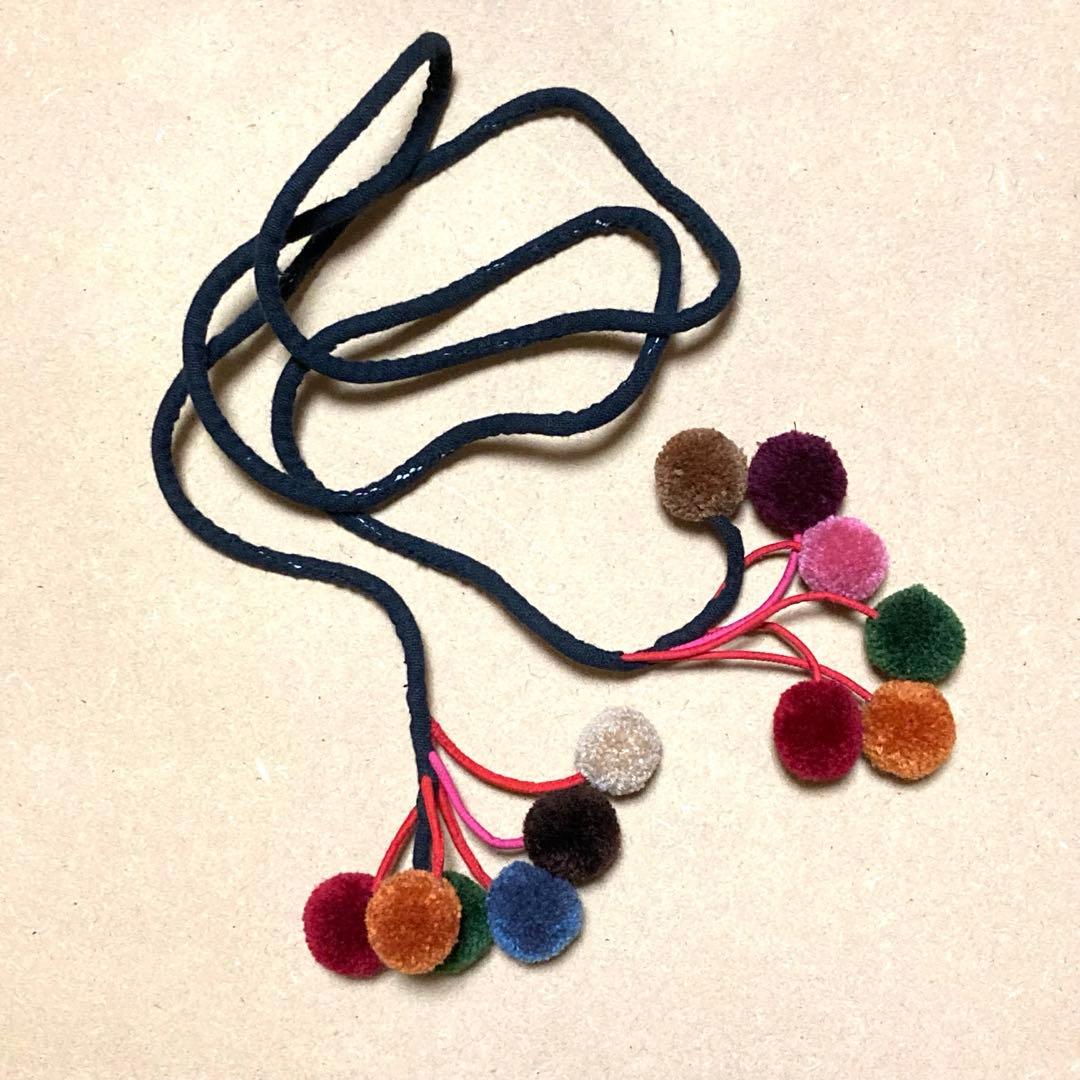 A polyester pompom belt with a dark cord and multiple colorful pom-poms attached at the ends on a light background.