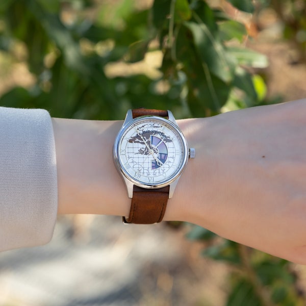 A silver-cased wristwatch with a brown leather strap and a white map-like dial is shown worn on a wrist outdoors.