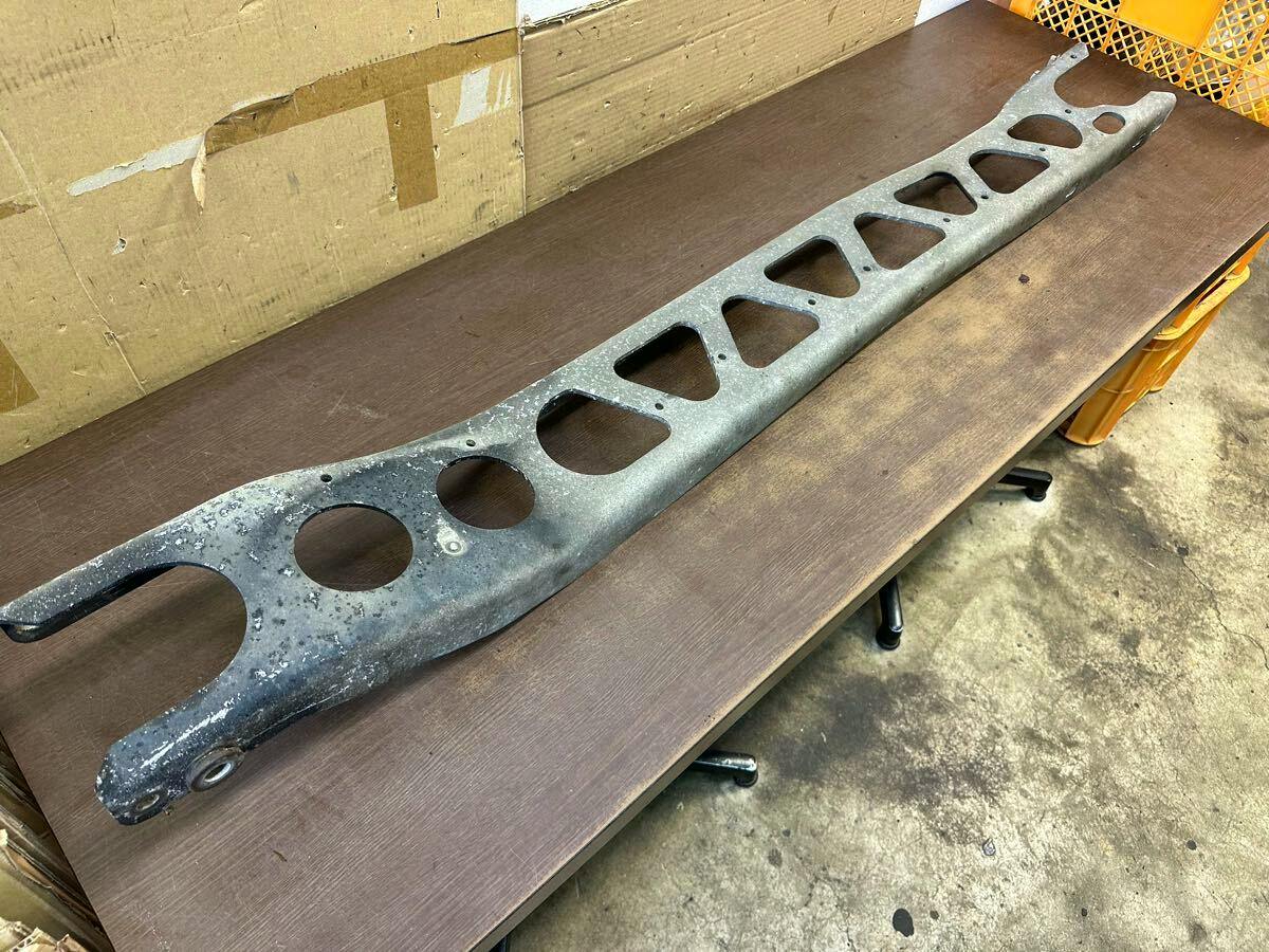 Long metal powerplant frame (car bracket) for an E-NA8C Roadster laid on a table, showing triangular and circular cutouts and surface corrosion.