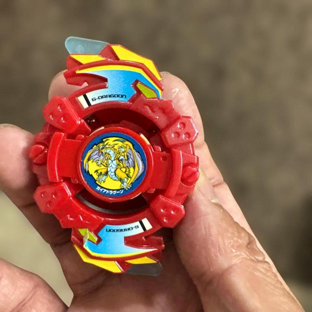 Hand holding the red Gaia Dragoon toy top showing the stickered face and colored fins.