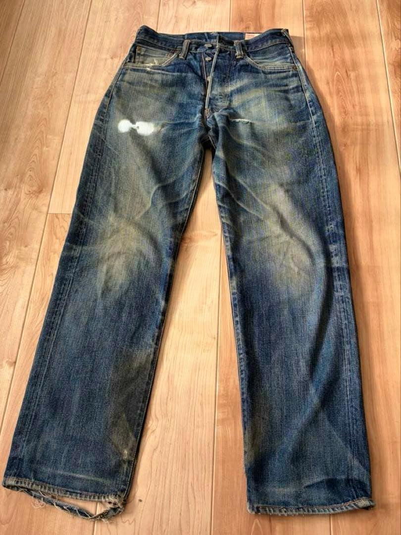 Evisu men's straight denim jeans laid flat on a wooden floor showing the front with fading, a small hole, and frayed hems.