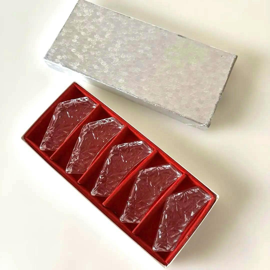 A boxed set of five clear glass chopstick rests arranged in a red-lined box with the silver lid placed beside it.