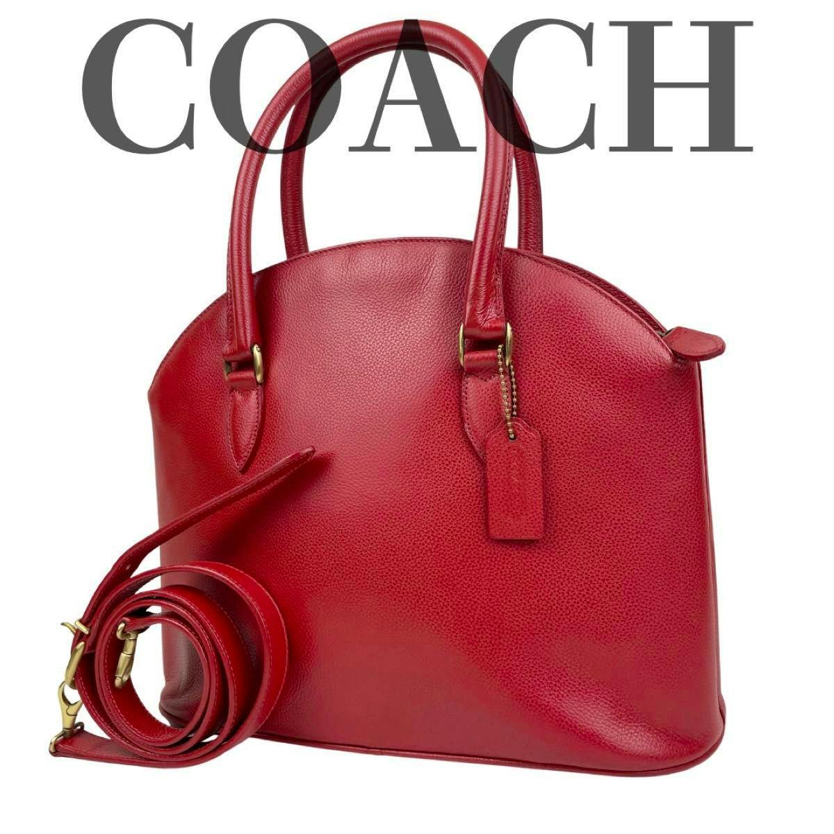 Red dome-shaped leather Coach handbag with top handles and a coiled detachable shoulder strap on a white background.