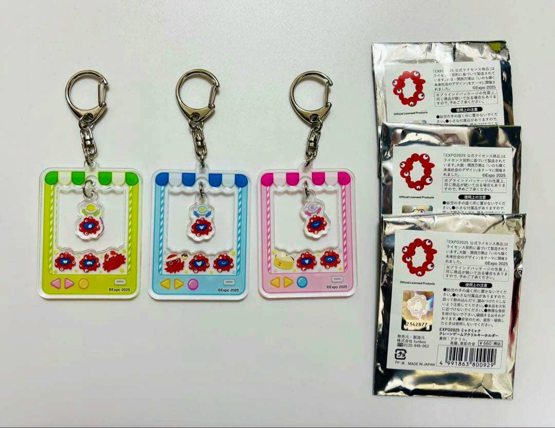Three EXPO2025 acrylic crane-game keychains (yellow, blue, and pink frames) shown with their foil packaging on the right.