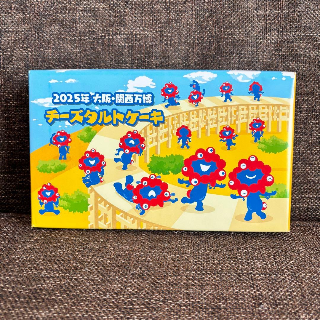 Front of the packaged box for the 2025 Osaka Expo cheese tart cake (7 pieces) showing colorful mascot characters on a blue and yellow background.