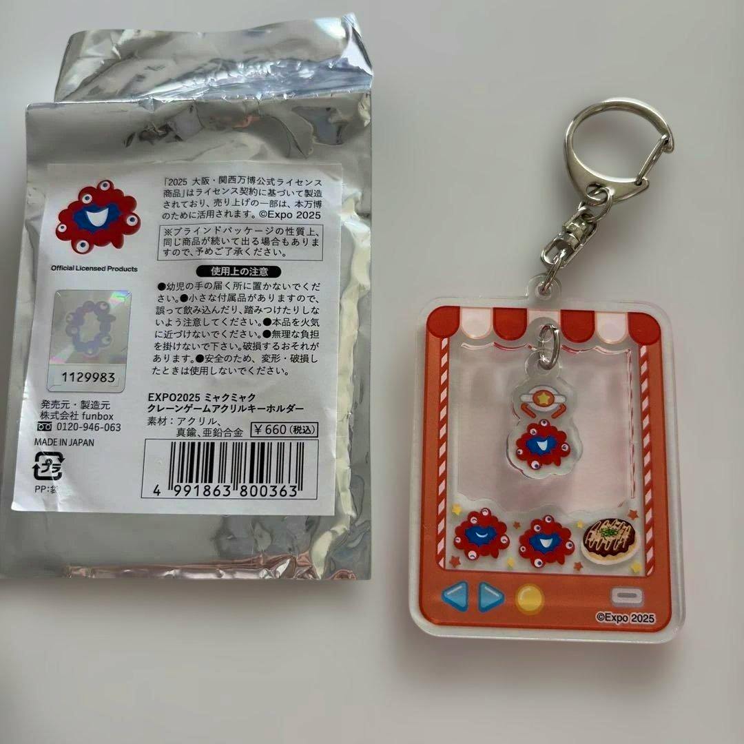 Crane-game themed acrylic EXPO2025 keychain showing a small red ミャクミャク charm inside a miniature claw-machine design, with silver foil packaging beside it.