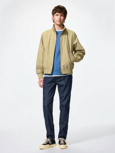 Male model wearing men's dark blue jeans with a beige jacket and sneakers, standing against a white background.