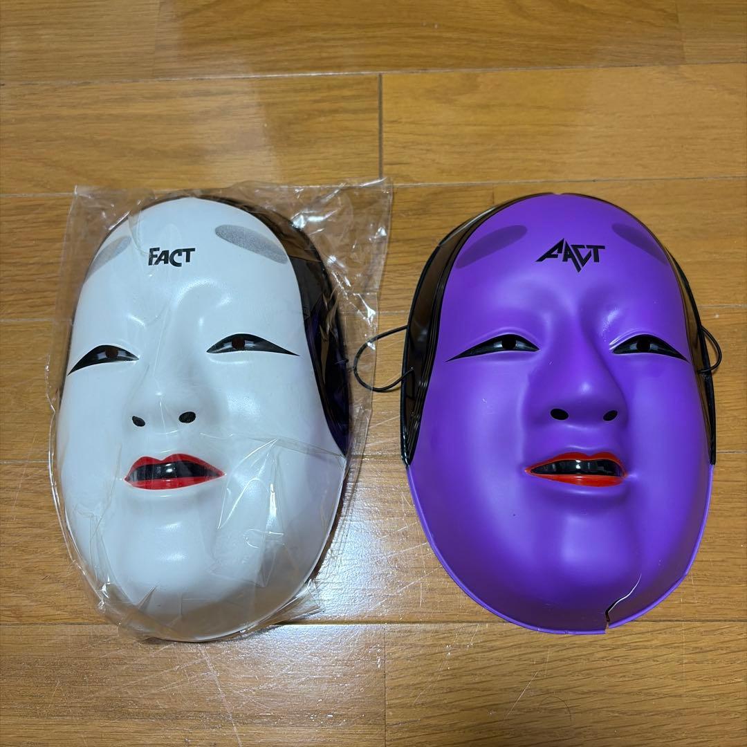 Two FACT plastic character (Noh-style) masks — a white mask still in plastic packaging and a purple mask — laid on a wooden floor.