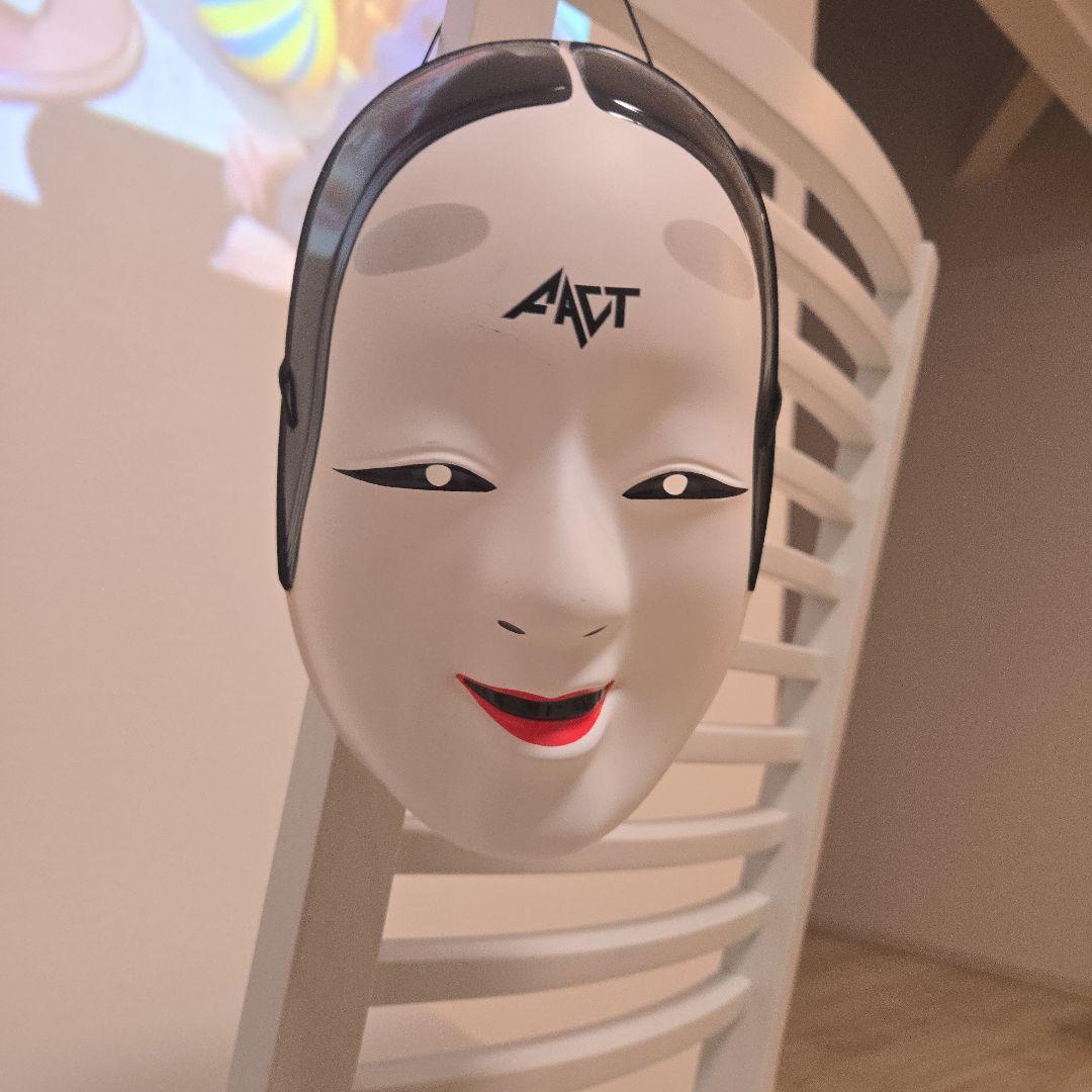 Front view of the FACT 能面 character plastic mask with black hair detailing, a FACT logo on the forehead, and red-painted lips hanging on a chair.