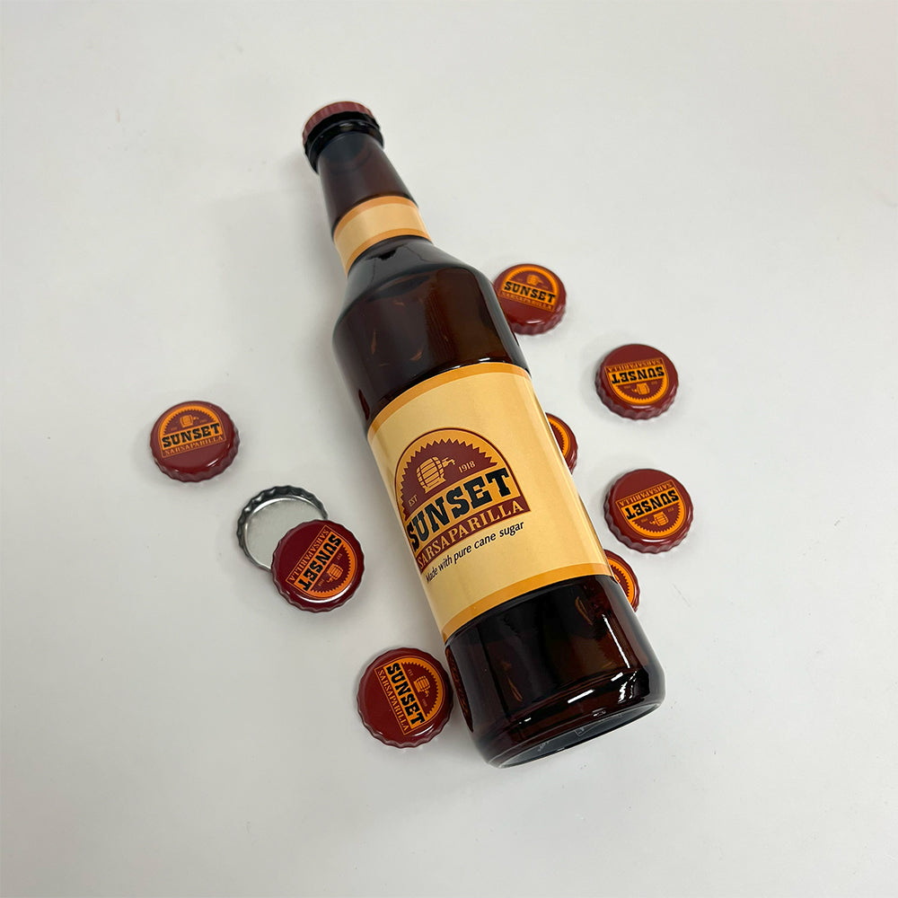 A Sunset Sarsaparilla glass bottle lying on a white background surrounded by several Sunset-branded bottle caps.