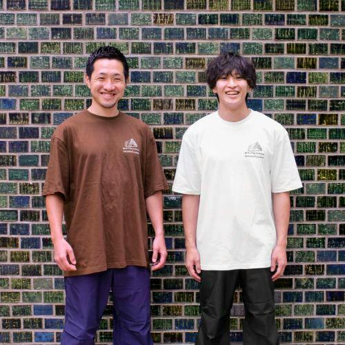 Two men wearing FANTASISTA CLIMBING collaboration cotton T-shirts (brown and white) standing against a tiled wall showing the front of the shirts.