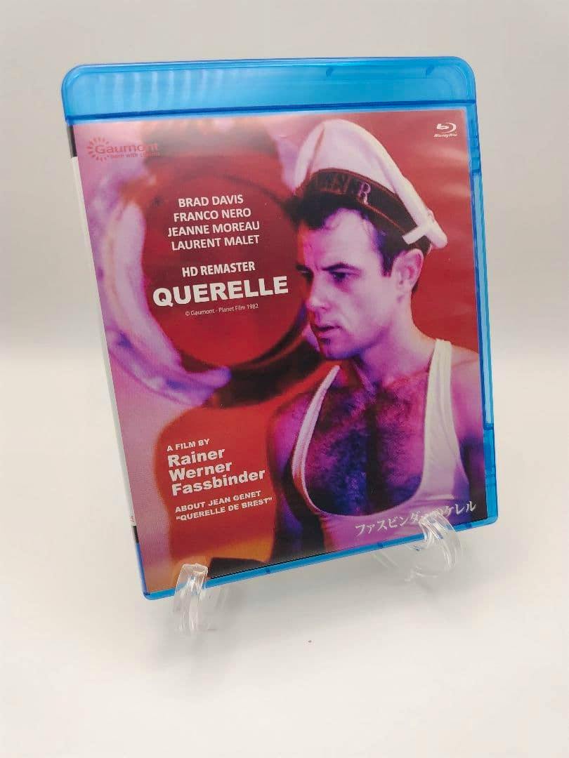 Blu-ray case of the film "Querelle" displayed on a clear stand, showing the cover art of a man in a sailor cap and tank top.