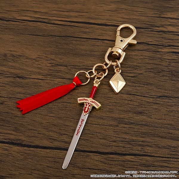 A miniature Saber weapon keychain charm from Fate/strange Fake with a small sword, red tassel, and gold clasp displayed on a wooden surface.
