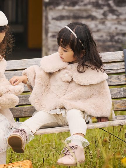 A child seated on a bench wearing a light beige faux-fur flower-motif poncho.