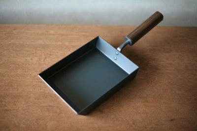 Rectangular tamagoyaki pan with wooden handle pictured on a wooden surface.