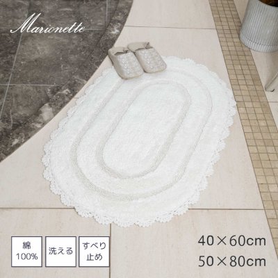 Oval ivory floor mat with scalloped frill edges placed on a tiled floor with a pair of slippers nearby.