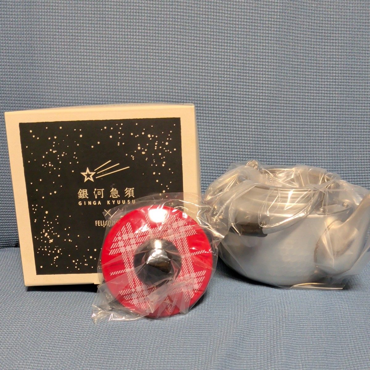 A stainless steel kettle with its box and a red lid knob, all wrapped in plastic, on a blue fabric surface.