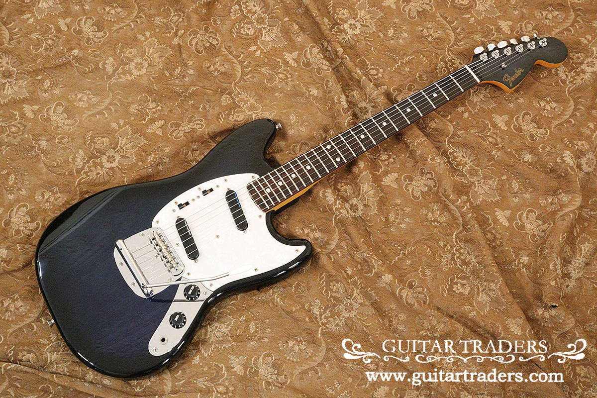 Oblique photo of a Fender Japan 1990's MG Custom Order electric guitar lying on a patterned fabric background, showing the body, neck, and headstock.