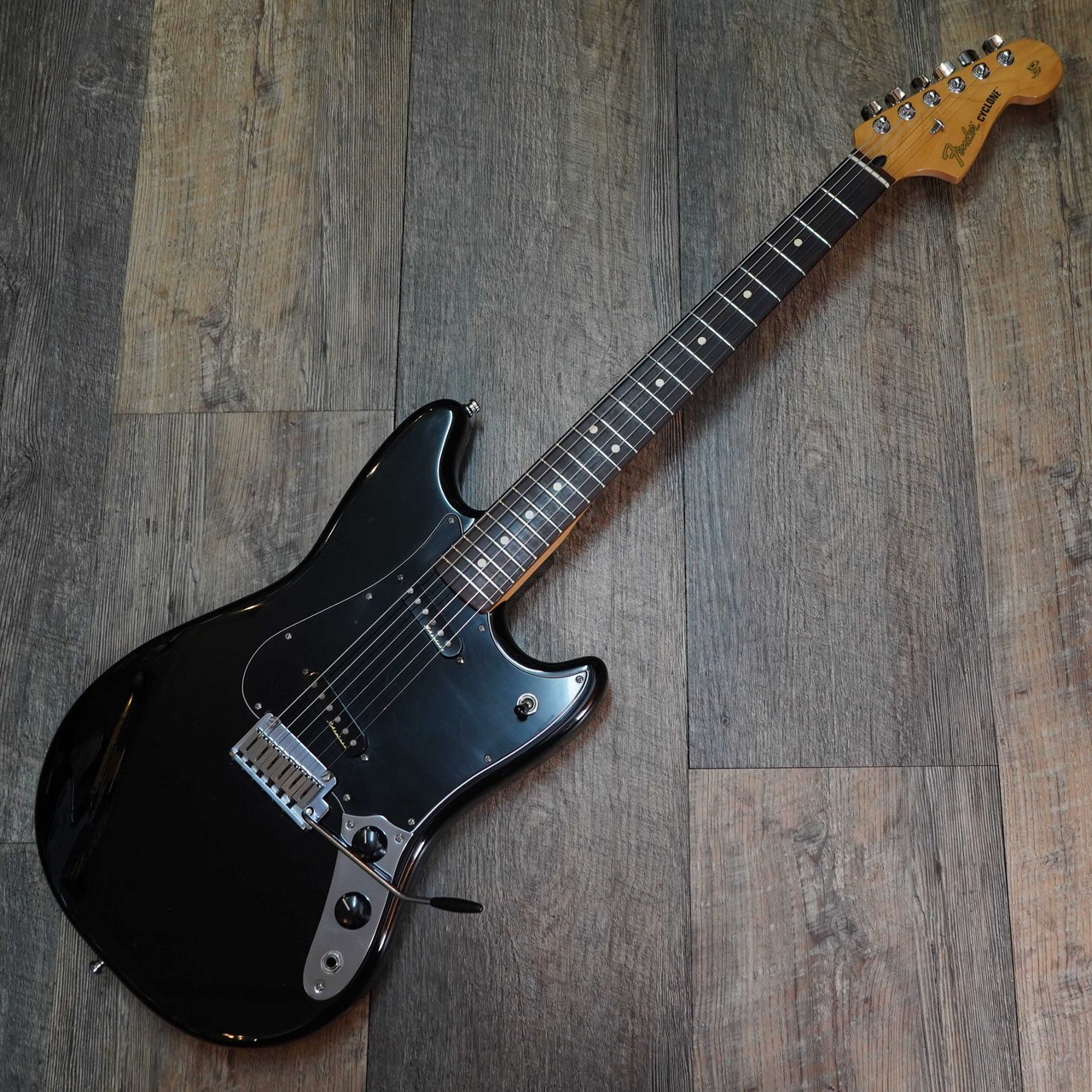 Black Fender Cyclone electric guitar with maple neck lying on a wooden floor.
