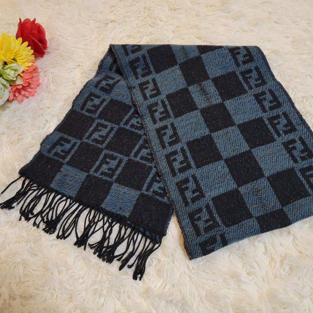 Blue and black checkered knit scarf with Fendi FF motif and fringed ends laid on a cream fur surface.
