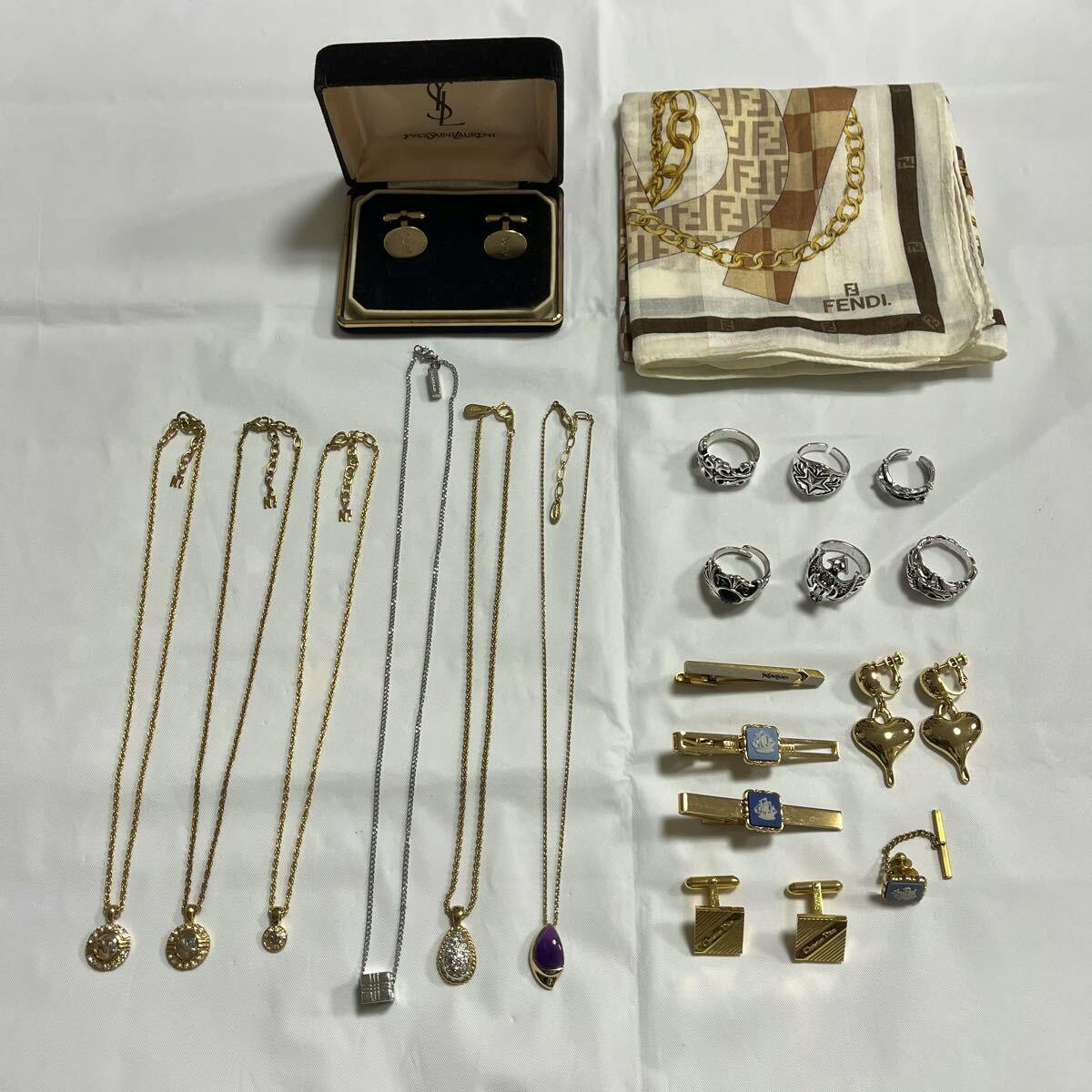 Flat lay of multiple necklaces with pendants, several rings, earrings, cufflinks and a folded Fendi scarf on a white background.