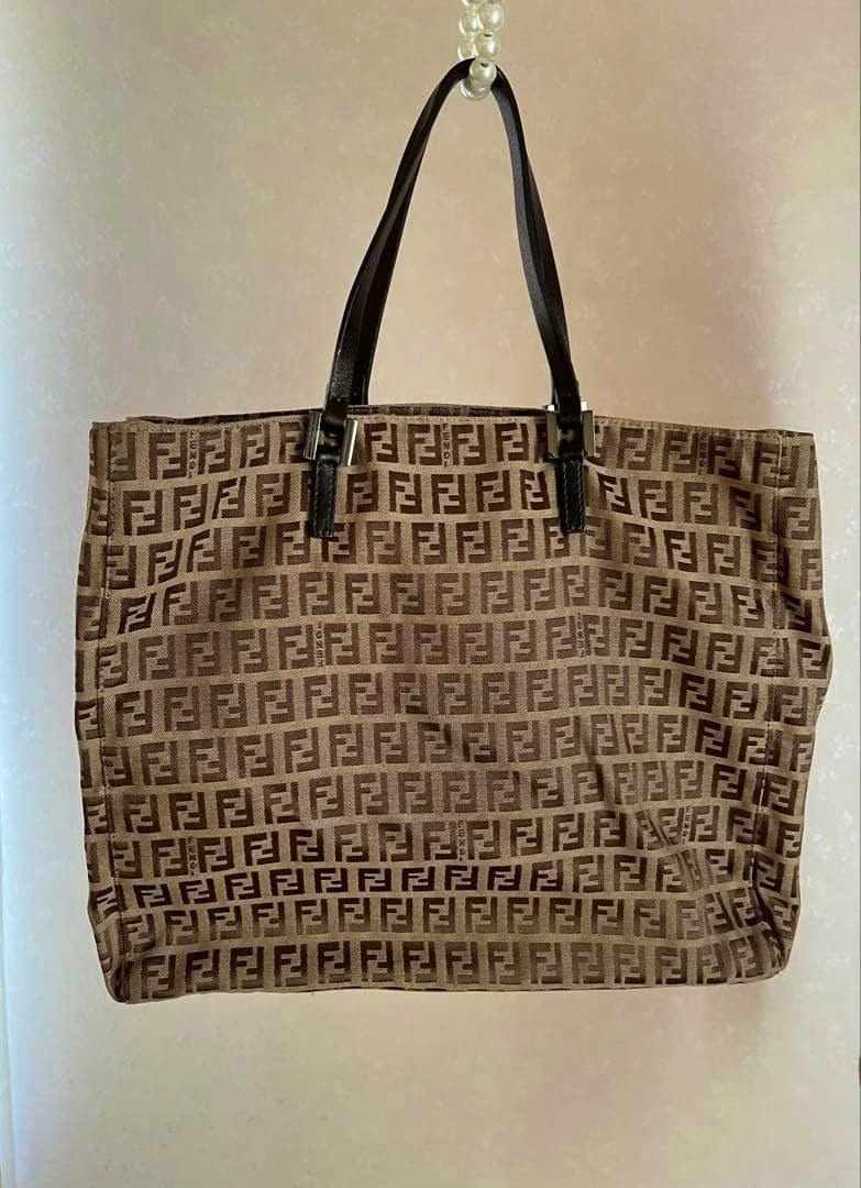 Brown FENDI tote handbag with repeating FF monogram and black shoulder straps hanging against a plain light background.