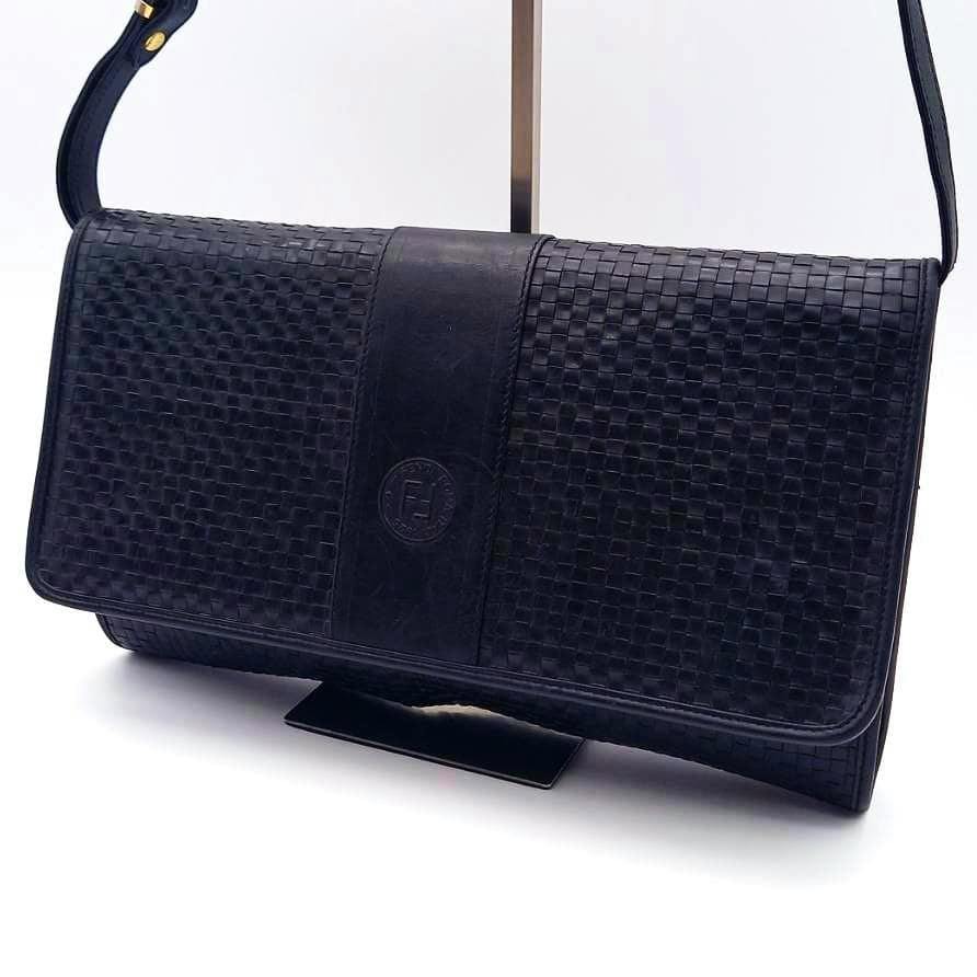 Black FENDI intrecciato cow leather shoulder bag with flap, central smooth leather panel and embossed logo, shown hanging from its strap on a stand.