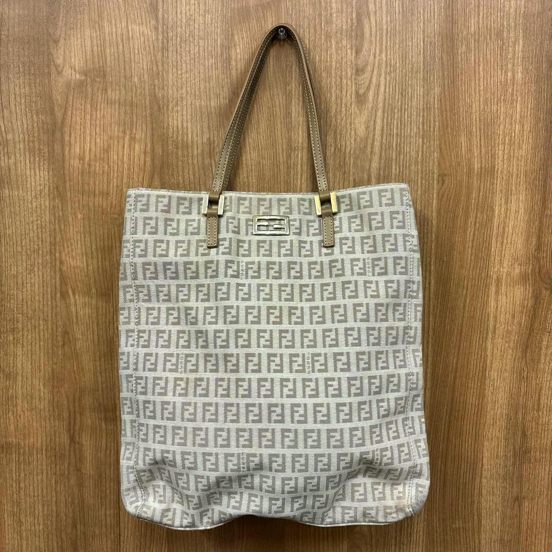 Beige FENDI Zuccino tote bag with repeating FF monogram pattern hanging against a wooden background.