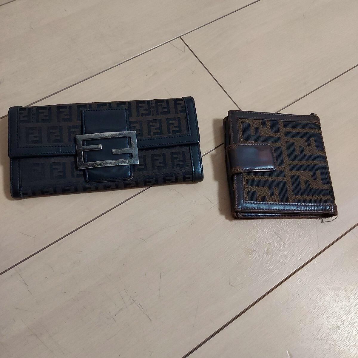 Two FENDI canvas wallets with Zucca monogram lying on a light wood floor.