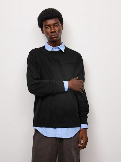 Male model wearing a black crew-neck sweater layered over a light blue button-up shirt against a plain background.