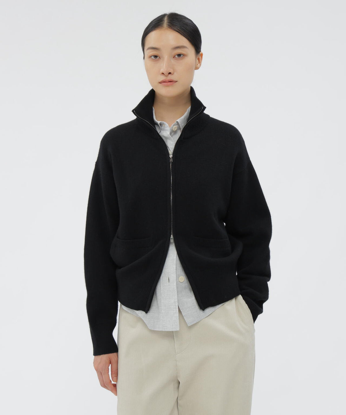 Front view of a model wearing a black fine wool loopback zip-up knitwear with patch pockets over a light button-up shirt.